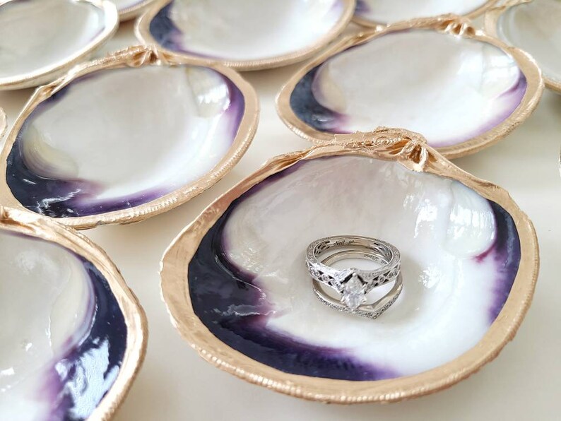 May include: A set of six white seashell ring dishes with gold trim and purple interiors. One dish has a silver ring with a diamond in the centre.