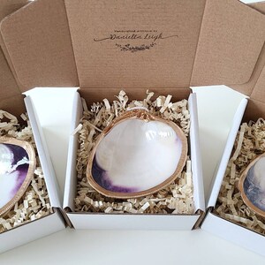 May include: Three white clam shell trinket dishes with gold trim and a purple interior are displayed in individual white boxes with brown paper packing material. Each box has a brown cardboard lid with the text "Handcrafted with love by Daniella Leigh" printed on the top.