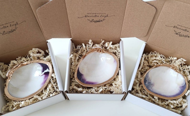 May include: Three white clam shell trinket dishes with gold trim and a purple interior are displayed in individual white boxes with brown paper packing material. Each box has a brown cardboard lid with the text "Handcrafted with love by Daniella Leigh" printed on the top.