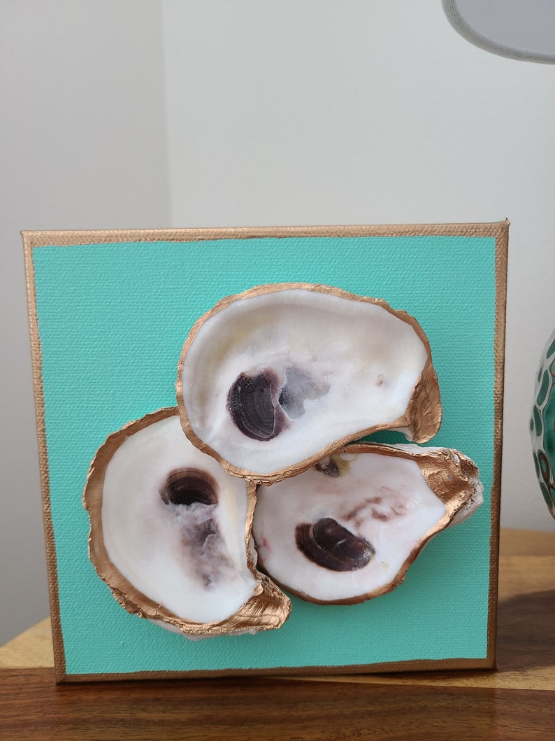 May include: Three oyster shells painted gold on a turquoise canvas. The shells are arranged in a triangular shape with the largest shell at the top.