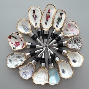 May include: A circular arrangement of twelve bottle stoppers featuring oyster shells as decorative tops. Each shell is edged in gold and displays unique patterns including nutcracker figures, holiday greenery, floral motifs, and solid pearlescent finishes.