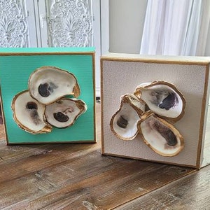 May include: Four square canvases with gold frames, each featuring a cluster of three oyster shells painted gold with a natural interior. The canvases are painted in shades of blue, green, and white.