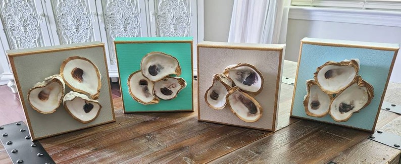 May include: Four square canvases with gold frames, each featuring a cluster of three oyster shells painted gold with a natural interior. The canvases are painted in shades of blue, green, and white.
