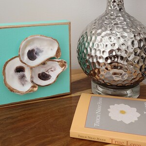 May include: A turquoise canvas with three oyster shells arranged in a triangular shape. The shells are painted gold and have a natural white interior. The canvas is framed with a gold border.