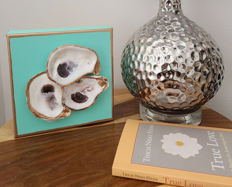 May include: A turquoise canvas with three oyster shells arranged in a triangular shape. The shells are painted gold and have a natural white interior. The canvas is framed with a gold border.