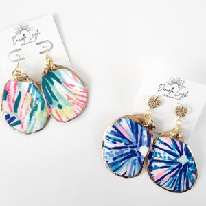 May include: Two pairs of dangle earrings featuring painted oyster shells with gold-colored edges. One pair has a colorful abstract design, and the other has a blue and pink pattern. The earrings are displayed on white cards with the text "Daniella Leigh Jewelry".