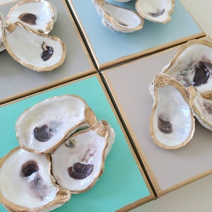 May include: Four square canvases with gold trim, each featuring a cluster of oyster shells with gold accents. The canvases are painted in shades of blue, green, and grey.