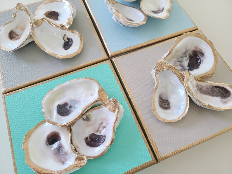 May include: Four square canvases with gold trim, each featuring a cluster of oyster shells with gold accents. The canvases are painted in shades of blue, green, and grey.