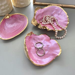 May include: Three decorative seashell dishes with a glossy pink interior and metallic gold-painted edges. One shell holds a string of white pearls, another holds two silver rings, and the third shell is empty. The shells are arranged on a surface.