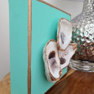 May include: A small square canvas painting with a teal background and gold trim. Three oyster shells are attached to the canvas, painted gold with a white interior.