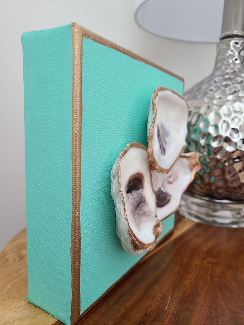 May include: A small square canvas painting with a teal background and gold trim. Three oyster shells are attached to the canvas, painted gold with a white interior.