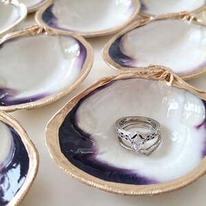 May include: A set of six clam shell ring dishes with gold trim. The shells are white with a purple iridescent sheen. One shell has a wedding ring set inside.