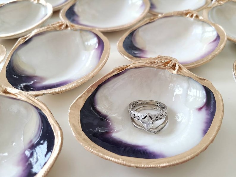 May include: A set of six clam shell ring dishes with gold trim. The shells are white with a purple iridescent sheen. One shell has a wedding ring set inside.