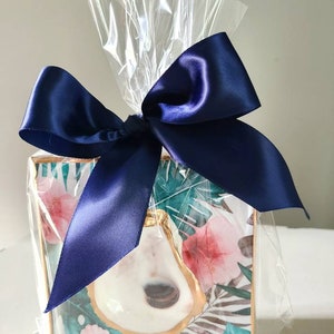 May include: A square, framed artwork with a tropical print featuring a white oyster shell with gold accents. The artwork is wrapped in clear cellophane and tied with a navy blue satin ribbon.