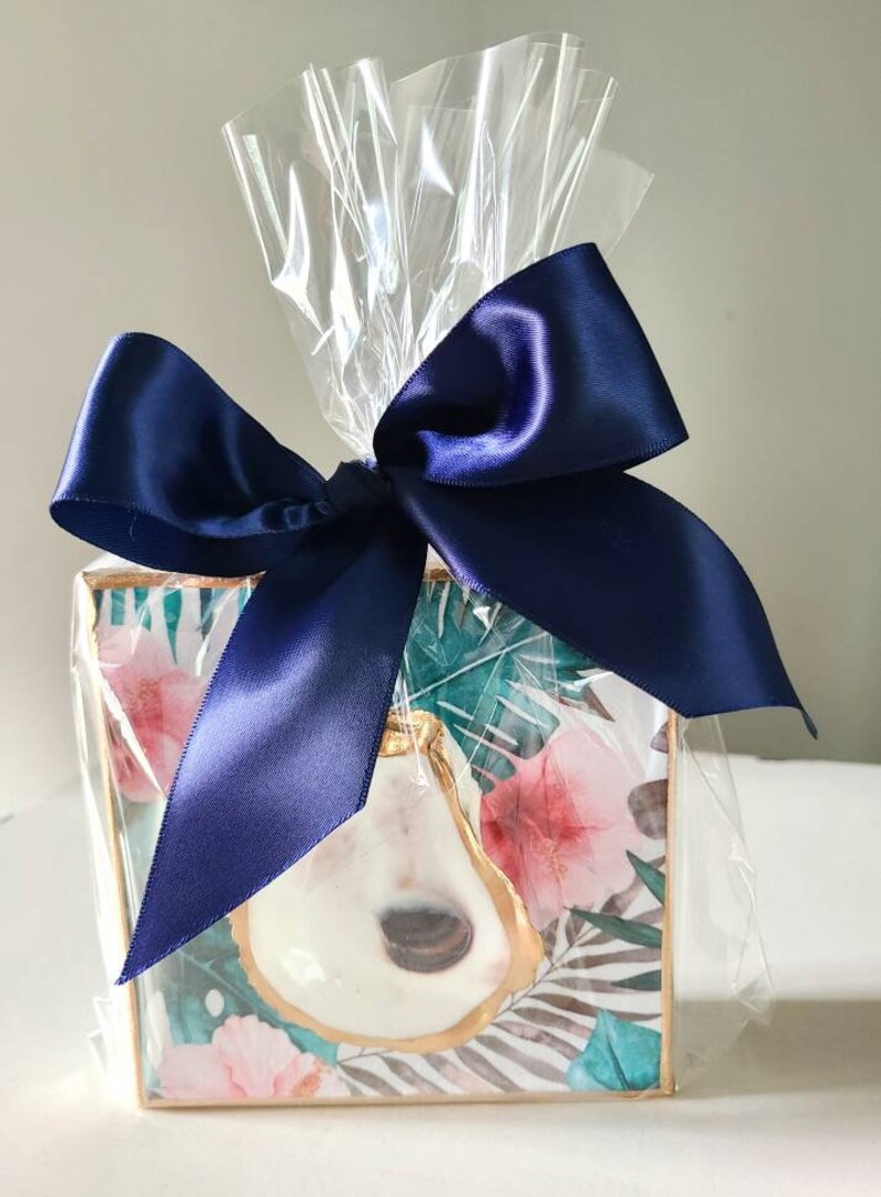 May include: A square, framed artwork with a tropical print featuring a white oyster shell with gold accents. The artwork is wrapped in clear cellophane and tied with a navy blue satin ribbon.