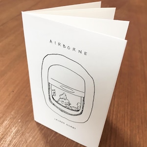 May include: A white card with black ink illustration of an airplane window with clouds outside. The text "AIRBORNE" is at the top of the card and "LAUREN NORBY" is at the bottom.