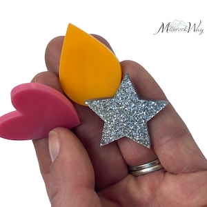 May include: A hand holding three decorative shapes: a pink heart, a yellow teardrop, and a silver glitter star. The shapes are smooth and have a simple, clean design. The background is white.