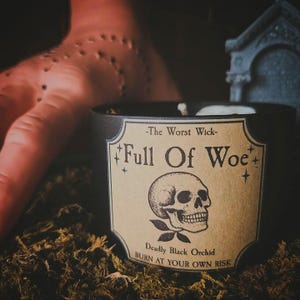 May include: A black candle with a label that reads "-The Worst Wick- Full Of Woe" and features a skull illustration. The label also states "Deadly Black Orchid BURN AT YOUR OWN RISK". The candle sits on a bed of green moss.