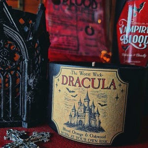 May include: A black candle in a cylindrical container with a beige label featuring a gothic castle illustration, bats, and the text The Worst Wick DRACULA Blood Orange & Oakwood BURN AT YOUR OWN RISK, positioned next to a black gothic lantern.