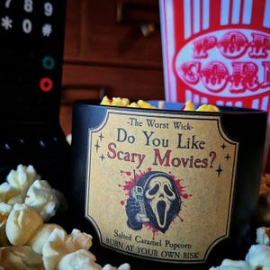 May include: A black candle container with a tan label featuring the text "The Worst Wick- Do You Like Scary Movies?" and "Salted Caramel Popcorn BURN AT YOUR OWN RISK" alongside an illustration of a masked figure holding a vintage telephone.