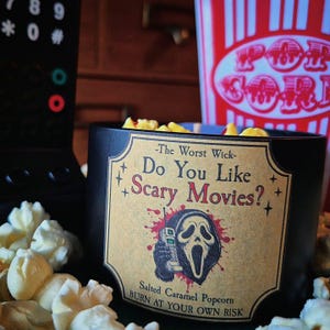May include: A black candle in a jar with a tan label featuring the text "The Worst Wick- Do You Like Scary Movies?" and an illustration of a masked figure holding a phone. The label also includes the text "Salted Caramel Popcorn" and "BURN AT YOUR OWN RISK".