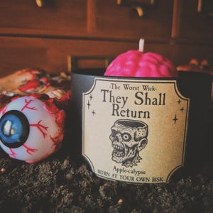 May include: A black candle holder with a pink brain-shaped candle. The label reads "They Shall Return" with a zombie illustration. An eyeball prop is visible on the left. The candle is apple-scented, as indicated on the label.