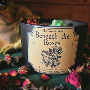 May include: A black candle with a label that reads "-The Worst Wick- Beneath the Roses." The label features a drawing of a frog and a rose. The candle is surrounded by dried rose petals. The text "Rose & Lavender for Luck BURN AT YOUR OWN RISK" is also on the label.
