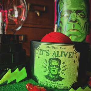May include: A black candle with a red wax top featuring a label that reads The Worst Wick IT'S ALIVE! Fearless Frankincense BURN AT YOUR OWN RISK, decorated with a green illustration of Frankenstein's monster and small star accents.