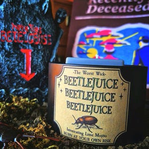 May include: A black candle with a light blue top and a label that reads "BEETLEJUICE" multiple times. The label also says "Intoxicating Lime Mojito" and "BURN AT YOUR OWN RISK". A tombstone and book are in the background.