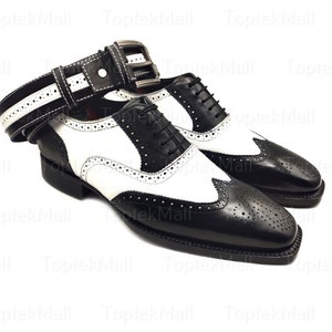 Può includere: Un paio di scarpe Oxford in pelle nera e bianca con un design a punta alare. Le scarpe hanno una punta in pelle bianca e un tallone in pelle nera. Le scarpe sono allacciate e hanno una cintura in pelle bianca con una fibbia nera.