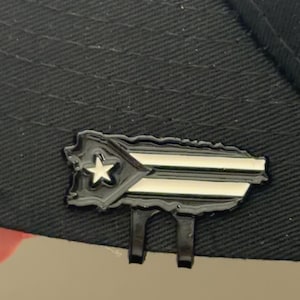 May include: A black hat with a Puerto Rican flag pin. The pin is a detailed outline of the island with a white star and horizontal stripes. The pin is black with white and silver accents.