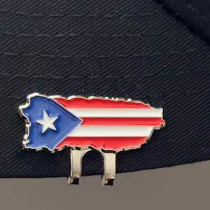 May include: A colorful pin in the shape of Puerto Rico's map, featuring the flag's design: a blue triangle with a white star, and red and white stripes. The pin is attached to a black hat.