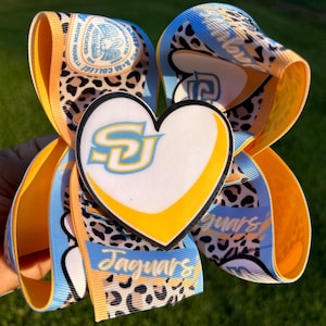 May include: A blue, yellow, and white hair bow with a leopard print design. The bow features a heart-shaped charm with the letters "SU" in white and a yellow outline. The bow also has the words "Jaguars" printed on it.