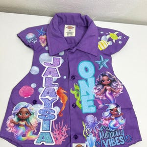 Op de afbeelding: Paarse button-down shirt met zeemeermingraphics en de tekst "Jalaysia ONE Mermaid Vibes". De shirt heeft illustraties van zeemeerminnen, zeedieren en bellen. De shirt heeft korte mouwen en een kraag.
