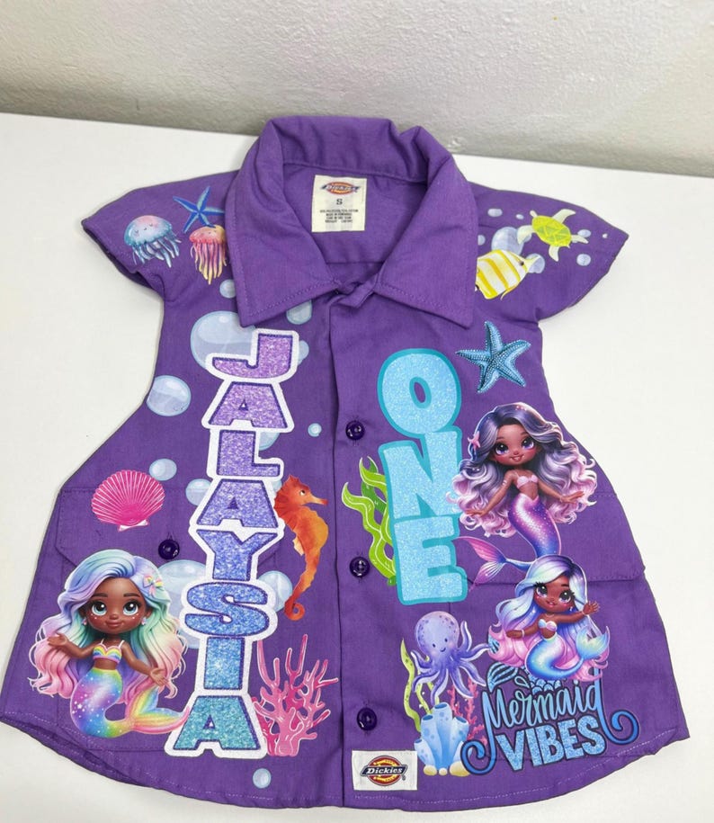 Op de afbeelding: Paarse button-down shirt met zeemeermingraphics en de tekst "Jalaysia ONE Mermaid Vibes". De shirt heeft illustraties van zeemeerminnen, zeedieren en bellen. De shirt heeft korte mouwen en een kraag.