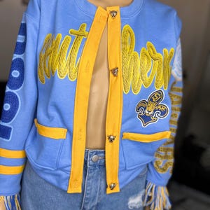 May include: A light blue jacket with yellow accents, featuring the embroidered text "Southern" in yellow script. The jacket has yellow trim, pockets, and decorative fringe. It is fastened with three decorative buttons.