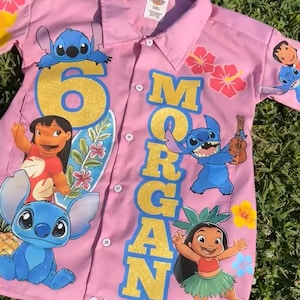 Op de afbeelding: Roze overhemd met knopen met personages uit de animatiefilm Lilo & Stitch. Het shirt heeft het nummer "6" en de naam "Morgan" in gouden glitterletters. Bloemen- en karakterafbeeldingen zijn op het shirt gedrukt.
