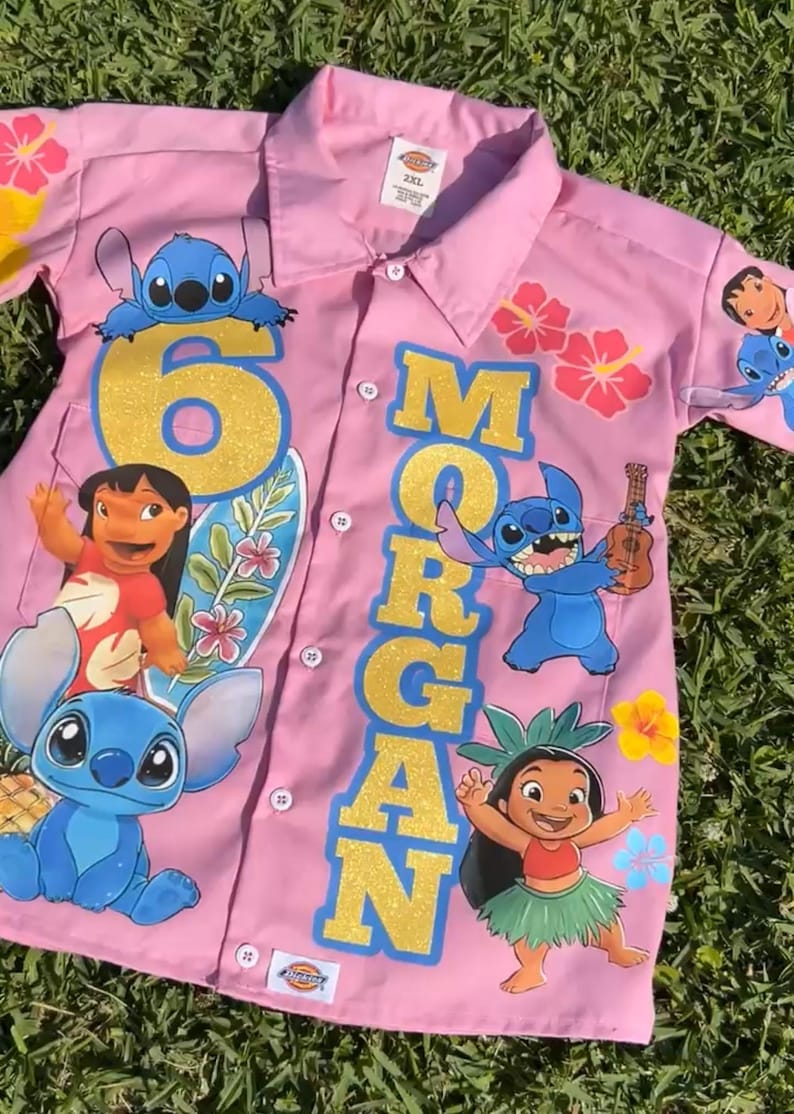 Op de afbeelding: Roze overhemd met knopen met personages uit de animatiefilm Lilo & Stitch. Het shirt heeft het nummer "6" en de naam "Morgan" in gouden glitterletters. Bloemen- en karakterafbeeldingen zijn op het shirt gedrukt.