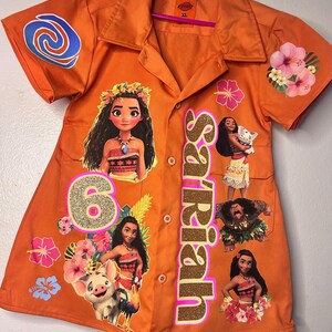 Op de afbeelding: Oranje overhemd met knopen met personages uit de animatiefilm Moana. Het shirt heeft een goudglitterende naam en het nummer 6. Bloemen- en wervelontwerpen zijn ook aanwezig.