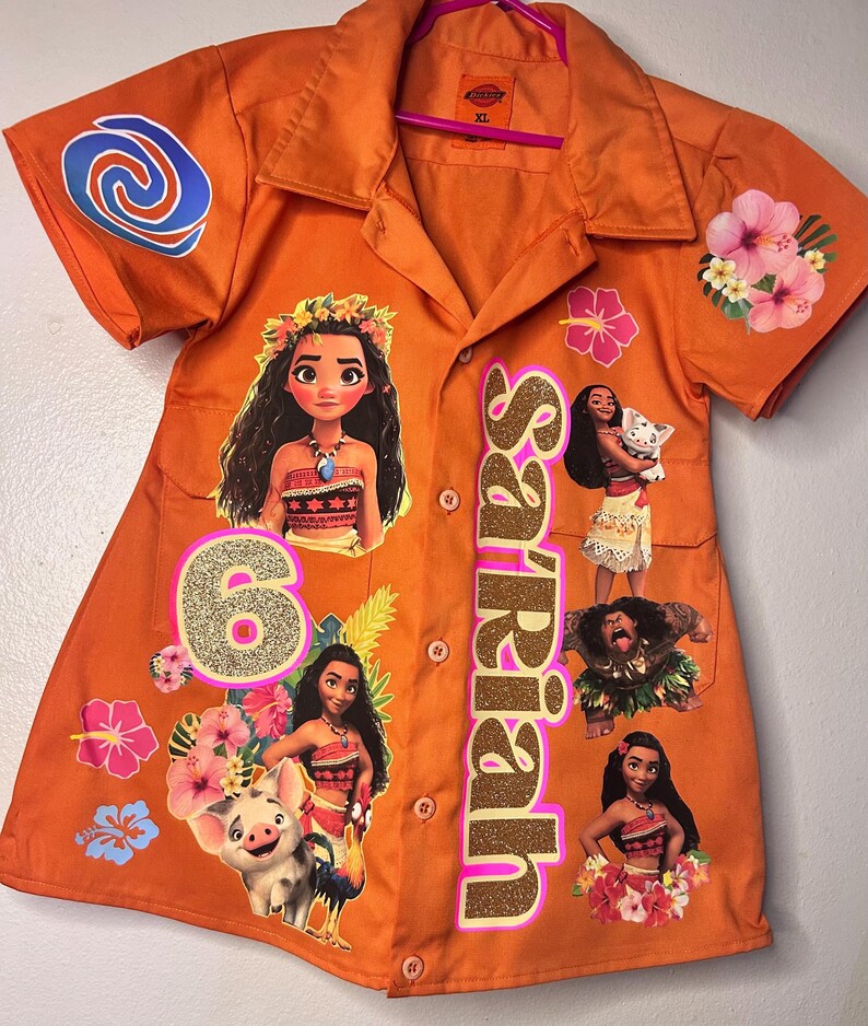 Op de afbeelding: Oranje overhemd met knopen met personages uit de animatiefilm Moana. Het shirt heeft een goudglitterende naam en het nummer 6. Bloemen- en wervelontwerpen zijn ook aanwezig.