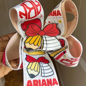 May include: A white hair bow made of grosgrain ribbon featuring a school-themed pattern with red pencils, rulers, and the name ARIANA printed in red. A central acrylic charm depicts a red bow tied around a yellow pencil and a white notebook.