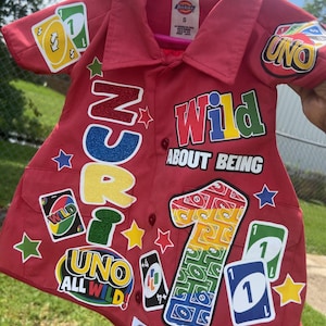 Op de afbeelding: Rood overhemd met knopen met het thema UNO-kaartspel. Voorzien van kleurrijke patches met de woorden "UNO", "Wild" en "1". Inclusief stervormige en kaartvormige patches. Het shirt hangt aan een roze hanger.