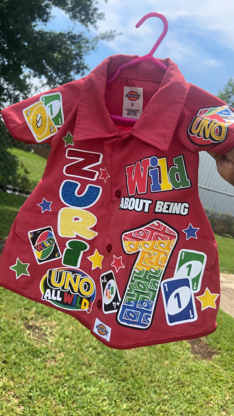 Op de afbeelding: Rood overhemd met knopen met het thema UNO-kaartspel. Voorzien van kleurrijke patches met de woorden "UNO", "Wild" en "1". Inclusief stervormige en kaartvormige patches. Het shirt hangt aan een roze hanger.