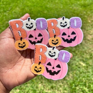 May include: Three Halloween decorations spelling "BOO". Each features a pink pumpkin with a carved face, a white pumpkin, and an orange pumpkin. The word "BOO" is in orange and purple, perfect for seasonal decor.