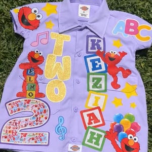 Op de afbeelding: Een lichtpaars overhemd met Elmo en Sesamstraat-thema's. Het shirt heeft het nummer "2" en het woord "TWO" in gouden glitter, samen met ABC-blokken en muzieknoten. Het shirt is versierd met sterren en Elmo-personages.