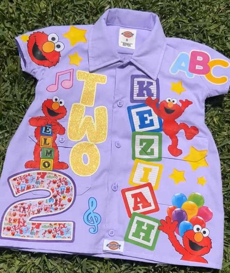 Op de afbeelding: Een lichtpaars overhemd met Elmo en Sesamstraat-thema's. Het shirt heeft het nummer "2" en het woord "TWO" in gouden glitter, samen met ABC-blokken en muzieknoten. Het shirt is versierd met sterren en Elmo-personages.