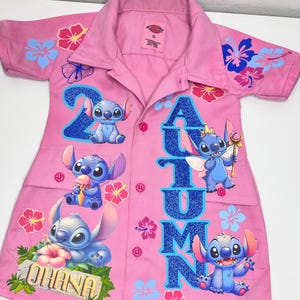 Op de afbeelding: Roze overhemd met Stitch-afbeeldingen en bloemmotieven. Het woord "Autumn" is geschreven in blauwe glitter. Het woord "Ohana" is ook zichtbaar.