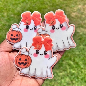 Puede incluir: Tres pegatinas de fantasmas de Halloween. Cada fantasma blanco tiene una cara sonriente, mejillas rosadas y un lazo naranja. Cada uno sostiene una calabaza. Las pegatinas se sostienen en una mano sobre un fondo verde.