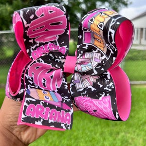 Peut inclure: Un grand nœud pour les cheveux fait à la main, avec un motif noir et blanc et des accents roses. Le nœud porte le nom "Ariana" et des graphiques de dessins animés colorés. Le nœud est tenu par une main.