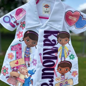 Op de afbeelding: Witte laboratoriumjas voor kinderen met Doc McStuffins-personage. De jas heeft een paarse glitter naam, karakterafbeeldingen en medische illustraties. Een roze hanger ondersteunt de jas.